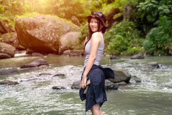 Genç Asyalı kadın gülümsüyor ve canlı yeşil bir ormanda arkasına bakıyor. Güneş şapkası ve açık hava kıyafetleri giyiyor. Yumuşak güneş ışığı altında..