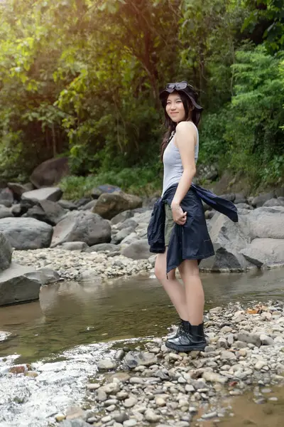 Yürüyüş kıyafetleri içinde genç bir Asyalı kadın ormanda bir nehirde kayaların üzerinde duruyor. Huzurlu bir doğa anının tadını çıkarırken gülümsüyor..