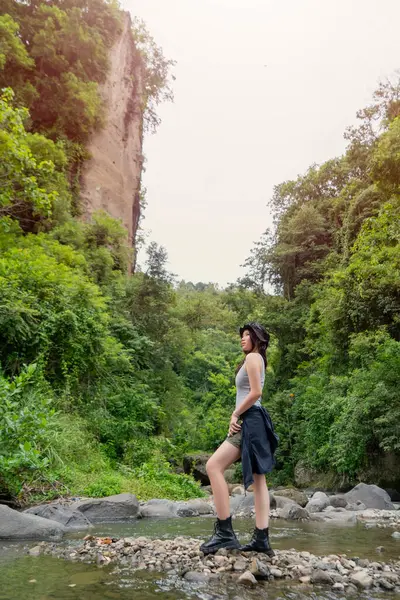 Genç Asyalı bir kadın, yoğun orman ve yüksek uçurumlarla çevrili kayalık bir nehir yatağında güvenle duruyor, doğanın tadını çıkarıyor.