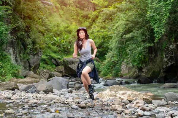 Neşeli genç Asyalı kadın canlı yeşilliklerle çevrili nehir taşlarının üzerinden koşarak doğa macerasının tadını çıkarıyor.