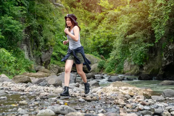 Neşeli genç Asyalı kadın canlı yeşilliklerle çevrili nehir taşlarının üzerinden koşarak doğa macerasının tadını çıkarıyor.