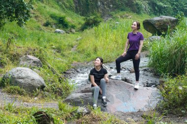 Spor kıyafetli iki Asyalı kadın nehir kenarında dinleniyor ve doğanın tadını çıkarıyorlar. Sağlıklı, sürdürülebilir yaşam tarzlarını yansıtıyorlar.