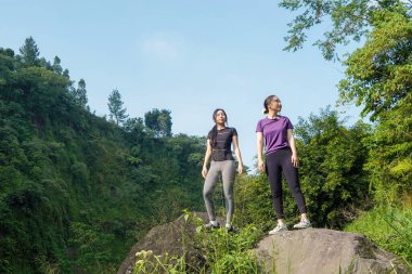 İki Asyalı kadın kayaların üzerinde dururlar. Çevreleri bereketli, sportif ve sürdürülebilir bir yaşam tarzını ön plana çıkarırlar..