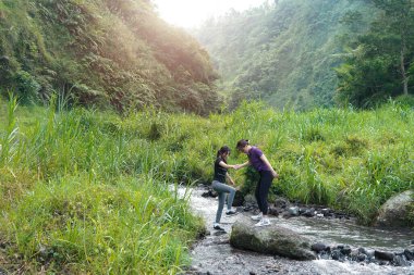 İki Asyalı kadın, bereketli bir dağ manzarasında açık hava egzersizi sırasında nehri geçerken birbirlerine destek oluyorlar.
