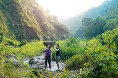 İki Asyalı kadın yemyeşil bir vadide kayaların üzerinde duruyor, açık hava egzersizlerini ve sürdürülebilir bir yaşam tarzını kucaklıyorlar..