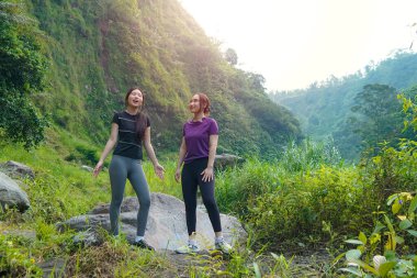 İki Asyalı kadın yemyeşil bir vadide kayaların üzerinde duruyor, açık hava egzersizlerini ve sürdürülebilir bir yaşam tarzını kucaklıyorlar..