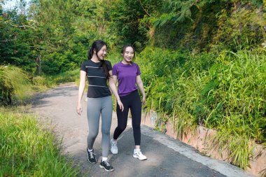 İki Asyalı kadın, doğa tarafından çevrelenmiş sağlıklı, aktif ve sürdürülebilir yaşam tarzlarını kucaklayarak, açık hava egzersizleri sırasında yemyeşil bir yolda yürürler..