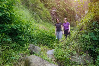 İki Asyalı kadın, doğa tarafından çevrelenmiş sağlıklı, aktif ve sürdürülebilir yaşam tarzlarını kucaklayarak, açık hava egzersizleri sırasında yemyeşil bir yolda yürürler..