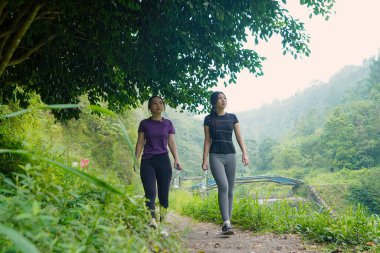 İki Asyalı kadın, doğa tarafından çevrelenmiş sağlıklı, aktif ve sürdürülebilir yaşam tarzlarını kucaklayarak, açık hava egzersizleri sırasında yemyeşil bir yolda yürürler..