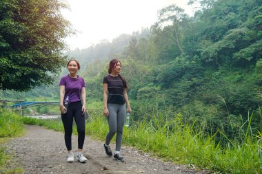 İki Asyalı kadın, doğa tarafından çevrelenmiş sağlıklı, aktif ve sürdürülebilir yaşam tarzlarını kucaklayarak, açık hava egzersizleri sırasında yemyeşil bir yolda yürürler..