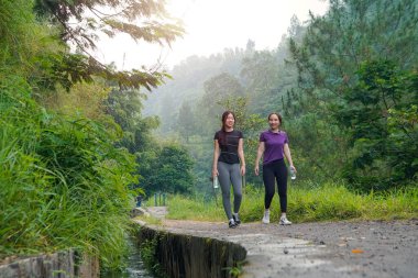 İki Asyalı kadın doğa yolunda yürüyor, açık hava egzersizlerinden ve yemyeşil bir ortamda sürdürülebilir yaşam tarzlarından keyif alıyorlar..