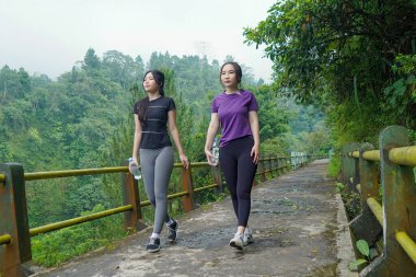 Spor giyimli iki Asyalı kadın ormanda bir köprüde yürüyor, dışarıda egzersiz, sağlıklı yaşam ve sürdürülebilir doğal yaşam tarzlarını teşvik ediyorlar..