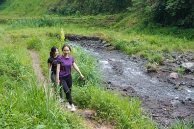 İki Asyalı kadın yemyeşil alanlarda yürüyüş yapıyor, doğa ile sağlıklı ve sürdürülebilir bir yaşam tarzını birleştiriyor..