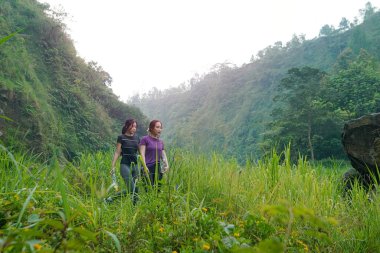 İki Asyalı kadın dağlarla çevrili yeşil bir vadide yürüyüş yapıyor. Egzersiz, doğa bağlantısı ve sürdürülebilir, sağlıklı yaşam tarzlarını destekliyorlar..