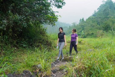 İki Asyalı kadın dağlarla çevrili yeşil bir vadide yürüyüş yapıyor. Egzersiz, doğa bağlantısı ve sürdürülebilir, sağlıklı yaşam tarzlarını destekliyorlar..