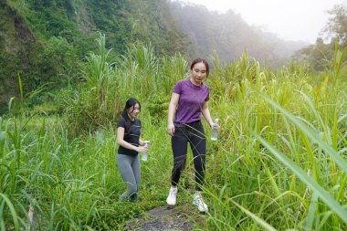 İki Asyalı kadın dağlarla çevrili yeşil bir vadide yürüyüş yapıyor. Egzersiz, doğa bağlantısı ve sürdürülebilir, sağlıklı yaşam tarzlarını destekliyorlar..