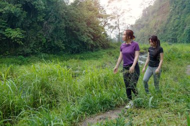 İki Asyalı kadın dağlarla çevrili yeşil bir vadide yürüyüş yapıyor. Egzersiz, doğa bağlantısı ve sürdürülebilir, sağlıklı yaşam tarzlarını destekliyorlar..