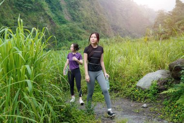 İki Asyalı kadın dağlarla çevrili yeşil bir vadide yürüyüş yapıyor. Egzersiz, doğa bağlantısı ve sürdürülebilir, sağlıklı yaşam tarzlarını destekliyorlar..