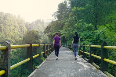 Manzaralı bir dağ köprüsünde egzersiz yapan iki Asyalı kadın sürdürülebilir sağlıklı yaşam tarzlarını destekliyor..