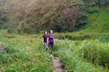 İki Asyalı kadın dağlarla çevrili yeşil bir vadide yürüyüş yapıyor. Egzersiz, doğa bağlantısı ve sürdürülebilir, sağlıklı yaşam tarzlarını destekliyorlar..