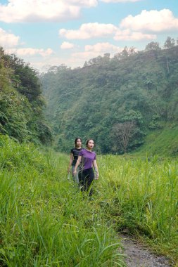 İki Asyalı kadın dağlarla çevrili yeşil bir vadide yürüyüş yapıyor. Egzersiz, doğa bağlantısı ve sürdürülebilir, sağlıklı yaşam tarzlarını destekliyorlar..
