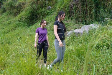 Doğa yolunda uzun otların arasında yürüyen iki Asyalı kadın egzersiz, spor ve sürdürülebilir yaşamı kucaklıyor.