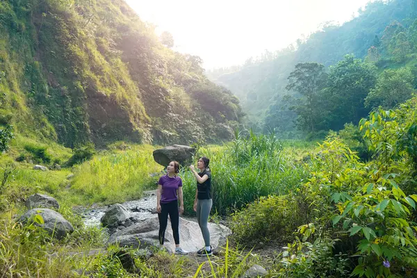 İki Asyalı kadın yemyeşil bir vadide kayaların üzerinde duruyor, açık hava egzersizlerini ve sürdürülebilir bir yaşam tarzını kucaklıyorlar..