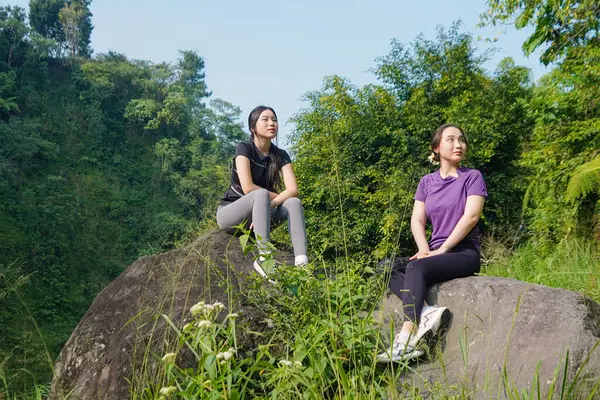 Spor giyim sektöründe iki Asyalı kadın açık hava egzersizlerinden sonra yemyeşil kayalıklarda rahat rahat dolaşır ve doğada sağlıklı ve sürdürülebilir bir yaşam tarzını benimserler..