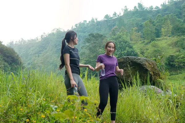 İki Asyalı kadın dağlarla çevrili yeşil bir vadide yürüyüş yapıyor. Egzersiz, doğa bağlantısı ve sürdürülebilir, sağlıklı yaşam tarzlarını destekliyorlar..