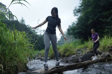 İki Asyalı kadın akarsuyu dikkatle geçerek düşmüş bir kütüğü kullanıyor, spordan ve sürdürülebilir yaşam tarzından zevk alıyorlar..