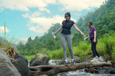 İki Asyalı kadın akarsuyu dikkatle geçerek düşmüş bir kütüğü kullanıyor, spordan ve sürdürülebilir yaşam tarzından zevk alıyorlar..