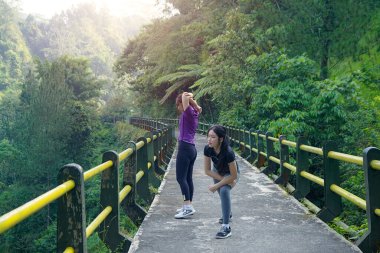 İki Asyalı kadın bir orman köprüsünde ısınma hareketleri yapıyor. Spor, doğa ve sürdürülebilir yaşam tarzlarını kucaklıyorlar..