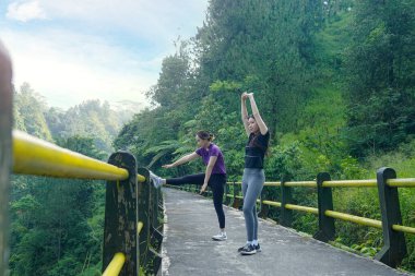 İki Asyalı kadın bir orman köprüsünde ısınma hareketleri yapıyor. Spor, doğa ve sürdürülebilir yaşam tarzlarını kucaklıyorlar..