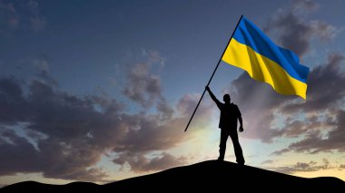 Küçük bir dağda duran bir adamın silueti, Ukrayna bayrağını sallıyor, arka planda şafak vakti bulutlu bir gökyüzü.