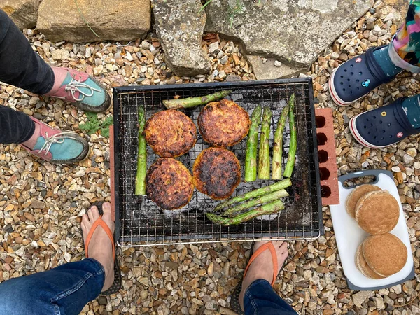 Three people around an outdoor portable barbecue cooking chicken cats burgers and fresh asparagus. High angle view point with feet in frame