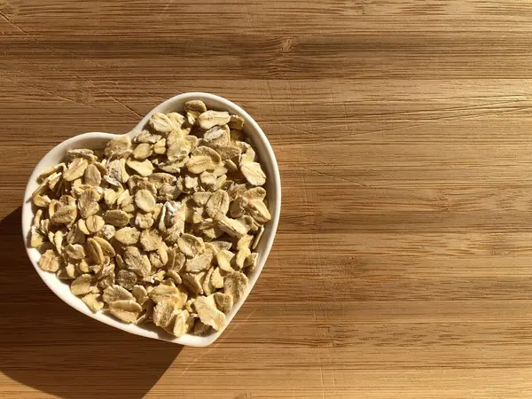 Healthy eating, healthy heart. Rolled oats in a heart shaped dish isolated on a wood background with copy space. Creative concept, diet and wellbeing