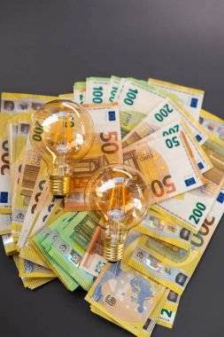 AB 'de elektrik maliyetleri. Elektrik faturalarının ödenmesi. Koyu gri arka plan üzerinde ampuller ve avro banknotları. Enerji sektöründeki kriz. Elektrik fiyatlarında artış. Elektrik faturalarının ödenmesi. 