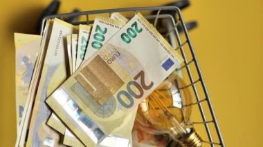 Elektrik fiyatları. Elektrik faturaları. Ampuller ayarlanmış ve avroluk banknotlar bir süpermarket arabasında sarı arka planda. Yukarıdan görüş. AB ülkelerinde elektrik maliyetleri düşük..