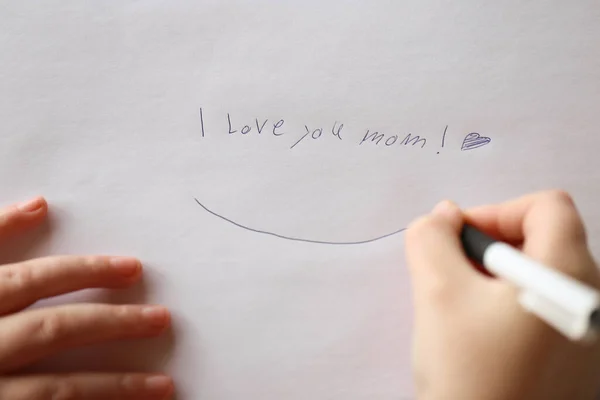 I love you mom inscription on a white sheet and a heart and a childs hand with a pen.Holiday of all mothers.Mothers day holiday.moms day concept.