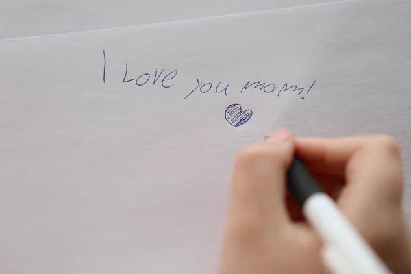 Mothers day holiday.moms day concept. I love you mom inscription on a white sheet and a heart and a childs hand with a pen.Holiday of all mothers