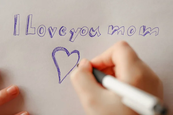 Mothers day holiday.moms day.I love you mom inscription on a white sheet and a heart and a childs hand with a pen.