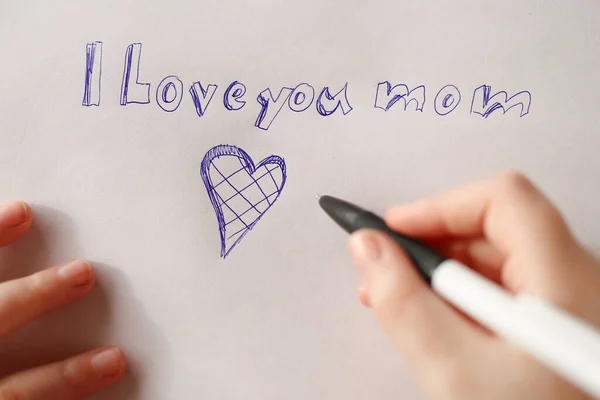 Mothers day holiday.moms day concept. I love you mom inscription on a white sheet and a heart and a childs hand with a pen.Holiday of all mothers