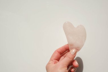 Gua sha facial massage device. gua sha scraper in the shape of a heart made of rose quartz in a female hand on a light background.beauty devices.Roller massager made of pink natural stone