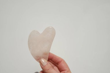 Gua sha facial massage . gua sha scraper in the shape of a heart made of rose quartz in a female hand on a light background.beauty devices.Roller massager made of pink natural stone