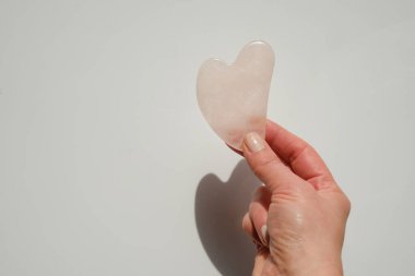 Gua sha facial massage device. gua sha scraper in the shape of a heart made of rose quartz in a female hand on a light background.beauty devices.Roller massager made of pink natural stone