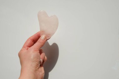  gua sha scraper in the shape of a heart made of rose quartz in a female hand on a light background.beauty devices.Roller massager made of pink natural stone