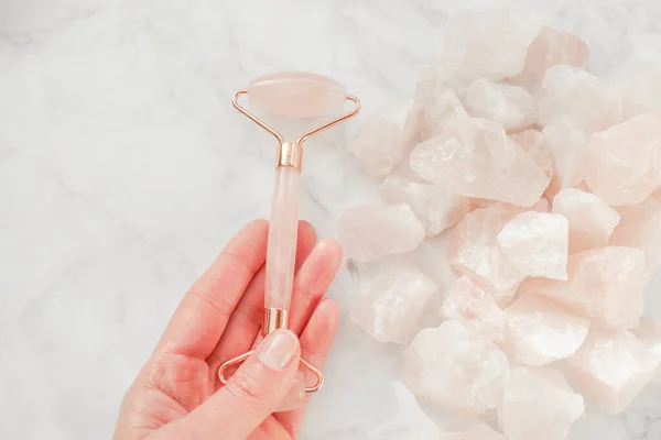 Rose quartz Massage roller close-up in a womans hand on rose quartz on gray marble background.facial massage device.gua sha roller made of pink natural stone