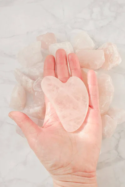Rose quartz massage scraper in the shape of a heart from natural stone on rose quartz on gray marble background.facial massage device.gua sha scraper.