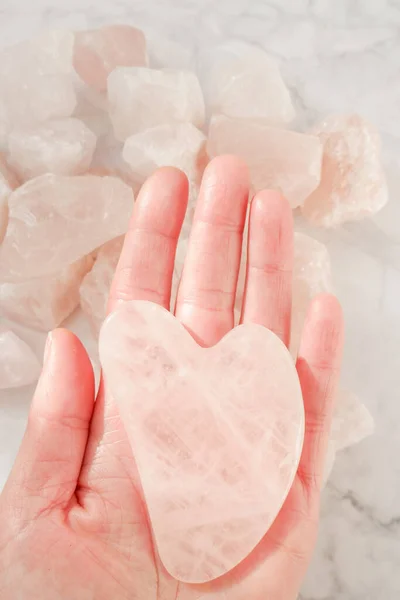 Rose quartz scraper in the shape of a heart from natural stone on rose quartz on gray marble background.facial massage device.
