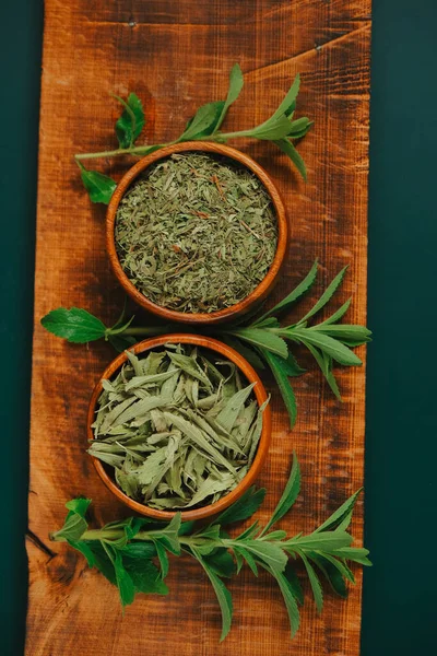 Stevia dry leaves and powder in round wooden bowls and fresh plant branches on a wooden board on a dark green background.sweet leaf sugar substitute.Dietary food sweetener.sweet plant.
